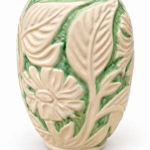 May include: A ceramic vase featuring a raised relief pattern of cream-coloured flowers and leaves against a recessed green glazed background. The cylindrical vessel has a smooth, glossy finish and a rounded shape with a wide opening at the top.