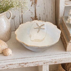 Peut inclure: Un bol en céramique blanc cassé à bords festonnés avec un motif d'étoile de mer. Le bol est posé sur une étagère blanche vieillie, à côté de coquillages, d'un vase de fleurs et d'un corail décoratif. Décor côtier.