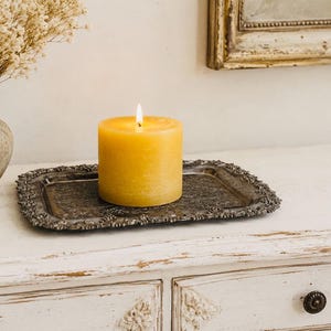 May include: A cylindrical yellow pillar candle with a lit wick, resting on a rectangular silver-toned metal tray with an ornate, textured border. The candle has a smooth, solid wax surface and emits a warm, steady flame in a vertical position.
