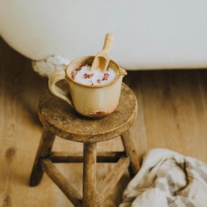 May include: A beige ceramic mug filled with bath salts and flower petals, with a small wooden scoop. The mug sits on a small, round, wooden stool. A white bathtub is in the background.