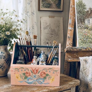 以下が含まれることがあります： 手描きの花柄と青いリボンが施された淡いピンク色の木製アート用品キャディ。キャディには木製の柄の絵筆が数本と、表面に絵具の跡が見える金属製の絵具チューブがいくつか収納されています。