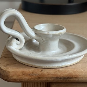 May include: Off-white ceramic candle holder with a circular base and a curved handle. The candle holder has a small cup to hold a candle and is sitting on a wooden surface. The design is simple and elegant.