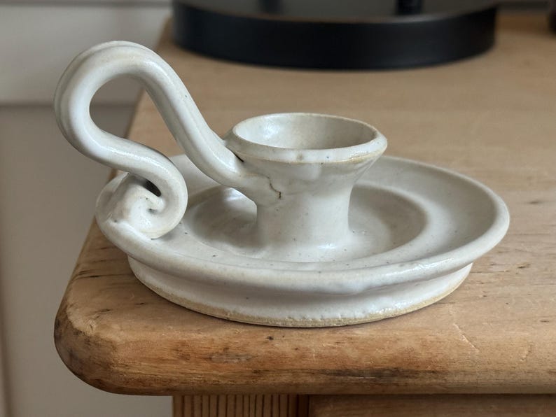 May include: Off-white ceramic candle holder with a circular base and a curved handle. The candle holder has a small cup to hold a candle and is sitting on a wooden surface. The design is simple and elegant.