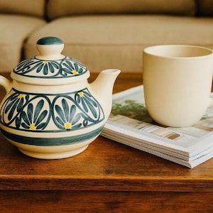 May include: A cream-coloured ceramic teapot with a matching lid, decorated with a blue and yellow floral pattern. A cream-coloured mug sits on a magazine nearby. The items are on a wooden table, suggesting a tea or coffee setting.