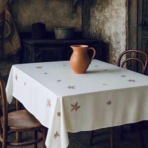 May include: A vintage kitchen setting with a white tablecloth adorned with floral embroidery, a terracotta jug, and wooden chairs. The room has aged walls and a dark wooden cabinet.