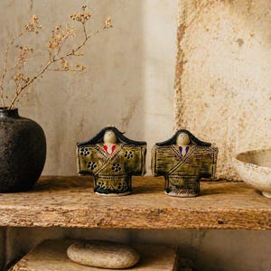 May include: Two ceramic dolls with kimono-style outfits in olive green and black, adorned with floral patterns. Each doll has a small face with a red and white detail. They sit on a rustic wooden shelf with other decorative items.