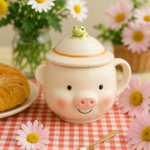 Könnte beinhalten: Eine skurrile Keramiktasse in Schweineform mit einem Deckel, der von einer kleinen grünen Froschfigur gekrönt wird. Die Tasse hat ein lächelndes Gesicht, rosa Wangen und einen Henkel. Die Tasse steht auf einer rot-weiß karierten Tischdecke, mit Blumen und einem Croissant.