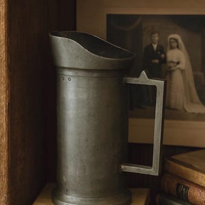 May include: A weathered pewter jug with a handle, sitting on a pile of aged books. The jug features a broad spout and a straightforward design. A framed monochrome photograph of a couple is visible in the background. The image conveys a sense of history and nostalgia.