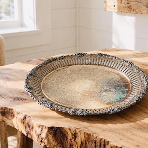 May include: A round, tarnished silver-tone metal tray featuring an intricate, embossed floral pattern in the centre. The tray has a raised, decorative border with a pierced, lattice-style rim and a textured, scalloped edge with dark patina accents.