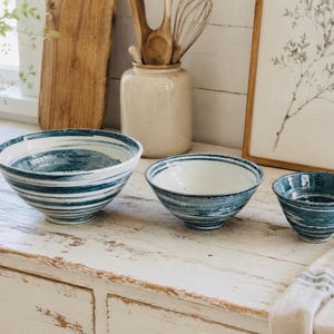 May include: Three ceramic bowls in varying sizes, each with a white interior and a blue and white striped exterior. The bowls are arranged on a distressed white wooden surface, suggesting a rustic or farmhouse aesthetic.