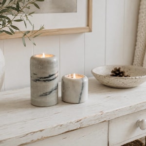May include: Two cylindrical marble candles with grey and black veining are lit, casting a warm glow. The taller candle stands next to a shorter, wider candle. Both feature smooth, polished stone surfaces with natural patterns in shades of white and grey.