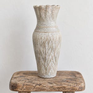 Peut inclure: Un grand vase cylindrique en céramique avec une finition texturée blanc cassé et beige. Le vase présente un bord festonné, un col étroit avec une bande horizontale et un corps décoré d'un motif en relief de fines lignes verticales.