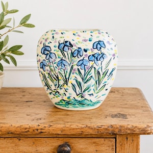 May include: A rounded ceramic vase with a cream-colored base featuring hand-painted blue flowers with green stems and leaves. The design includes scattered small purple and yellow brushstrokes across the surface, creating a textured floral pattern.