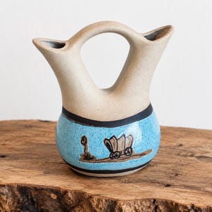 May include: A ceramic wedding vase with a double-spout design and a bridge handle. The upper portion is matte beige, while the lower bulbous section is bright blue with black speckles, featuring a hand-painted brown covered wagon illustration.