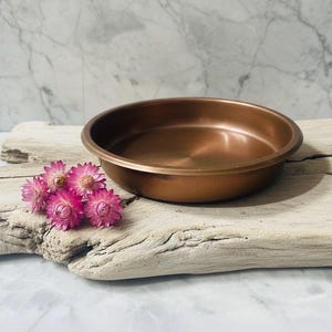 May include: A copper-coloured, shallow bowl with a rounded rim. The bowl sits on a piece of driftwood, with a cluster of pink flowers to the left. The background is a marble-patterned surface.