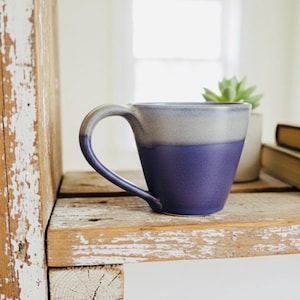 May include: A two-tone ceramic mug with a curved handle. The mug transitions from a light gray at the top to a deep purple at the bottom. It sits on a rustic wooden shelf with a small succulent and two vintage books.