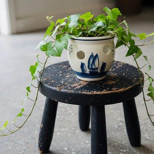 May include: A small, round, black wooden stool supports a white ceramic planter with a blue sailboat design. The planter is filled with green ivy, with vines cascading down the sides. The stool has three legs.