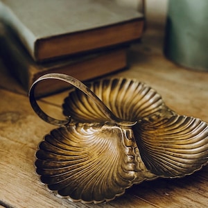 May include: A vintage, silver-toned serving dish with a shell-like design and a handle. The dish is placed on a wooden surface, with books and a wicker basket in the background. The dish has three compartments.