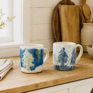 May include: Two ceramic mugs with hand-painted designs. The left mug features a light green pine tree against a blue background with a yellow circle. The right mug displays multiple dark blue pine trees painted in a textured style on a white base.