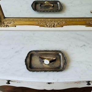 May include: A classic wristwatch with a brown leather strap and gold-coloured case sits on a silver tray. The tray is on a white marble surface, with a gold-framed mirror above. A second tray with a watch is reflected.