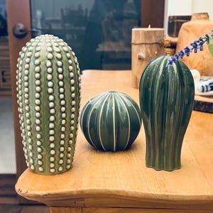 Peut inclure: Trois figurines de cactus en céramique dans des tons de vert variés. La plus grande est cylindrique avec des points blancs en relief, celle du milieu est une sphère avec des lignes verticales blanches, et la troisième est en forme de goutte.