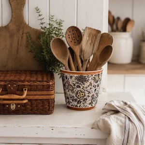 Peut inclure: Un pot à ustensiles en céramique orné d'un motif floral détaillé dans des tons de brun, vert et jaune, rempli d'une collection d'ustensiles de cuisine en bois comprenant des cuillères, une cuillère à trous et une spatule, avec un bord brun.