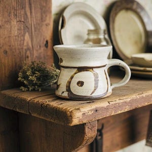 Könnte beinhalten: Eine handgefertigte Keramiktasse mit cremefarbener Glasur und braunem, abstraktem Design steht auf einem rustikalen Holzregal. Das Regal beherbergt auch Teller, eine Reibe und getrocknete Kräuter, was eine gemütliche, vintage Küchenästhetik erzeugt.