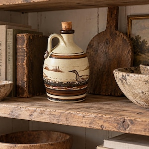 May include: A ceramic jug with a cream-coloured base, featuring a brown painted duck illustration and horizontal bands in shades of brown and black. The jug has a single handle, a narrow neck, and a cork stopper, with a small circular emblem on the handle.