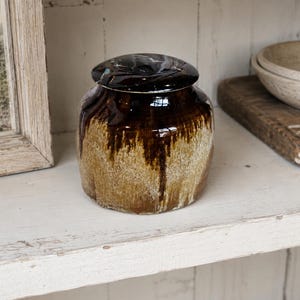 Peut inclure: Un petit pot en céramique rond avec couvercle, présentant une glaçure brun foncé brillante sur le dessus qui coule sur une base texturée dans des tons beige et brun clair. Le récipient a un corps large et légèrement conique avec un couvercle lisse.