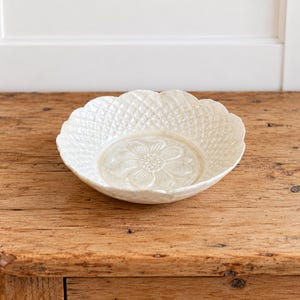 May include: A shallow, off-white ceramic bowl featuring a scalloped edge and a textured, diamond-patterned exterior. The interior base of the bowl displays a raised, embossed floral design with a central circular pattern and radiating petals.