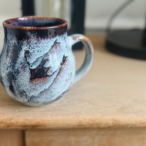 May include: A handmade ceramic mug with a unique glaze. The mug features a dark brown rim, transitioning to a light blue and brown mottled pattern on the body. It has a rounded shape with a sturdy handle, perfect for enjoying coffee or tea.