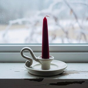 May include: A white ceramic candle holder with a red candle. The candle holder has a circular base and a curved handle. The candle is a deep red color and is unlit. The background is a window with a snowy scene.