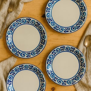 Può includere: Quattro piatti in ceramica bianca con un bordo decorativo blu con un motivo a spirale. I piatti sono disposti su una superficie di legno chiaro, con una forchetta e un cucchiaio visibili.