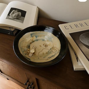 May include: A ceramic bowl with a black rim and a cream interior with blue details. Inside the bowl are a gold wristwatch, a gold necklace, and gold hoop earrings. The bowl sits on a dark wooden surface. Magazines and books are in the background.