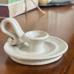 May include: Off-white ceramic candle holder with a circular base and a curved handle. The candle holder has a small cup to hold the candle and is placed on a wooden surface. The design is simple and elegant.