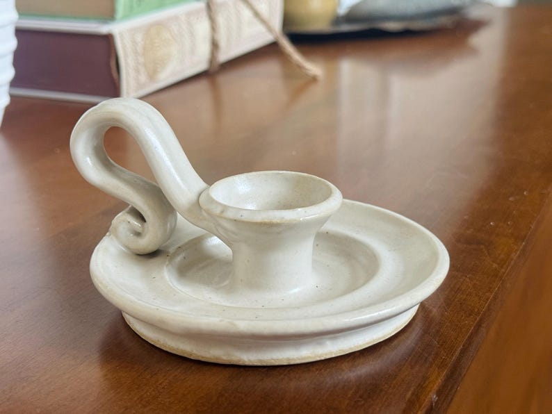 May include: Off-white ceramic candle holder with a circular base and a curved handle. The candle holder has a small cup to hold the candle and is placed on a wooden surface. The design is simple and elegant.