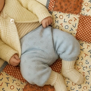 May include: A baby dressed in a cream cardigan, striped top, and light blue knitted trousers with white socks. The baby rests on a patchwork quilt with letters and floral designs.