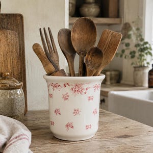 May include: A white ceramic utensil holder featuring a red floral and ribbon pattern, filled with various wooden kitchen tools including spoons, a fork, and a spatula. The cylindrical container has a smooth finish and a slightly flared top rim.