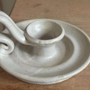 May include: Off-white ceramic candle holder with a circular base and a curved handle. The candle holder has a small cup to hold the candle and is made of speckled ceramic material. The design is simple and elegant, suitable for various home decor styles.