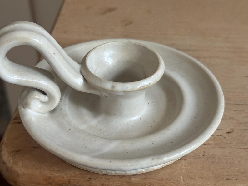 May include: Off-white ceramic candle holder with a circular base and a curved handle. The candle holder has a small cup to hold the candle and is made of speckled ceramic material. The design is simple and elegant, suitable for various home decor styles.
