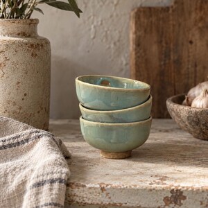 May include: A stack of three small, round ceramic bowls with a glossy, light seafoam green glaze and a visible, unglazed tan base. The bowls feature a slightly irregular, organic shape with smooth, curved edges and a reflective, glassy surface finish.
