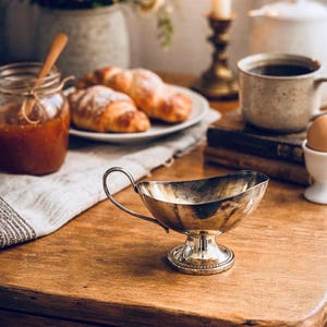 May include: A silver gravy boat with a handle sits on a wooden table, alongside croissants, coffee, and a jar of jam. A lit candle adds to the warm, inviting breakfast setting. The scene evokes a sense of comfort.