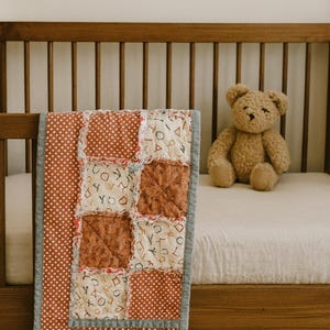 Puede incluir: Una colcha de bebé hecha a mano con un diseño de patchwork en tonos óxido, crema y blanco, con lunares y patrones de letras. Un oso de peluche marrón está sentado en la cama junto a la colcha. La colcha mide aproximadamente 91 x 122 cm.