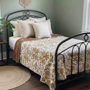 Op de afbeelding: Een slaapkamer met een smeedijzeren bed, versierd met een bloemenquilt en crèmekleurige kussens. Een macramé wandkleed en een rond jute vloerkleed zijn zichtbaar. De muren zijn geschilderd in een gedempt groen.