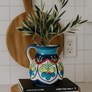 Puede incluir: Una jarra de cerámica azul y blanca con un diseño floral, que contiene ramas verdes y frondosas. La jarra se encuentra sobre una pila de libros en una bandeja de madera, con una tabla de cortar de madera en el fondo.