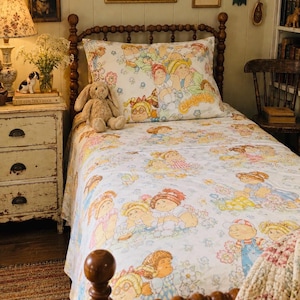 May include: A twin-sized bed featuring a white sheet set and pillowcase decorated with a repeating pattern of Cabbage Patch Kids characters in various outfits. A tan plush rabbit sits on the bed, which has a dark brown wooden spindle headboard.