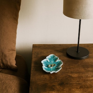 May include: A teal leaf-shaped ceramic dish holds two gold rings on a dark wooden nightstand. A brown pillow and a lamp with a beige shade are in the background. The lamp has a black base and stem.