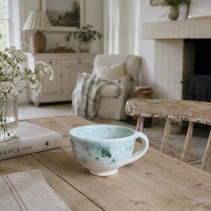 Op de afbeelding: Een lichtblauwe en witte keramische mok met een handvat staat op een houten tafel. De mok heeft een gespikkeld ontwerp. Een heldere glazen vaas met witte bloemen en een boek met de titel "THE KINFOLK HOME" staan ook op de tafel. De kamer heeft een neutraal kleurenpalet.