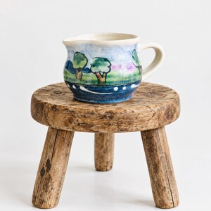 Könnte beinhalten: Ein Keramik-Milchkännchen mit einer handgemalten Landschaftsszene aus grünen Bäumen, blauem Himmel und einem dunkelblauen Boden mit weißen Punkten. Das Kännchen hat einen geschwungenen Henkel und einen kleinen Ausguss mit glänzender Oberfläche.