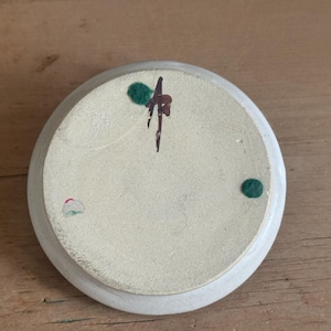May include: A small, off-white ceramic dish with a slightly raised edge. The underside features a speckled texture and is decorated with a dark brown and green abstract design, along with two green and one pink dot.
